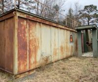 Rusty metal container in overgrown outdoor area.